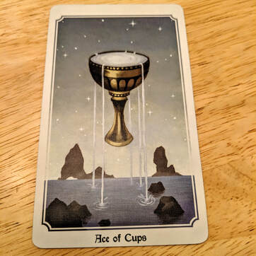 Picture of the Ace of Cups tarot card. Card depicts an overflowing goblet. Water flows out of the cup into the sea below, against a backdrop of stars.