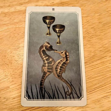 Picture of the Two of Cups tarot card. Card depicts two entwined seahorses beneath two upright goblets.
